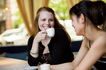 Two women are sat at a table smiling at each other, one women is facing the camera holding a mug, the other is facing away from the camera